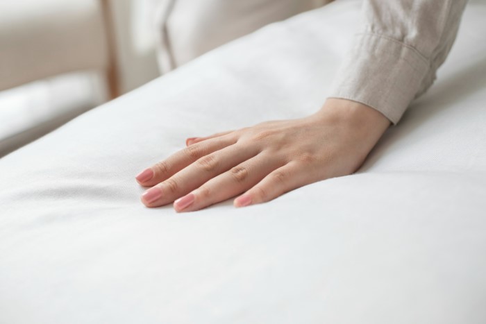 Detalhe mostrando uma mão feminina tocando a superfície macia de um colchão coberto com lençol branco, e que afunda levemente.