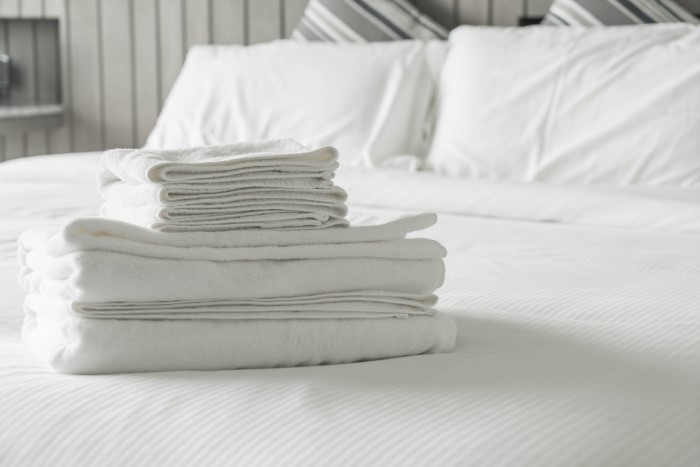 Detalhe mostrando toalhas brancas empilhadas sobre cama de casal com lençol e travesseiros brancos.