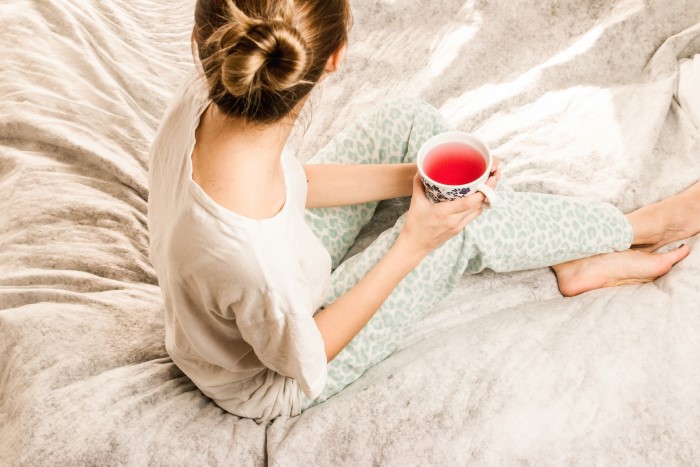 Mulher loira com coque no cabelo, vista de costas e de cima, sentada sobre a cama. Ela veste um pijama e segura uma xícara de chá. O edredom é branco com estampa mármore.