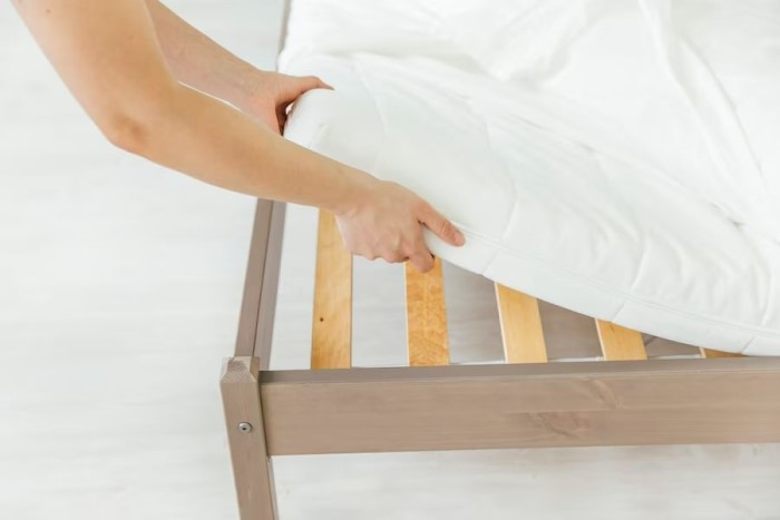 Imagem mostrando mãos femininas de pele clara levantando um colchão branco de cima de uma tradicional com base de estrado de ripas.  A cama é de madeira. O piso é claro.