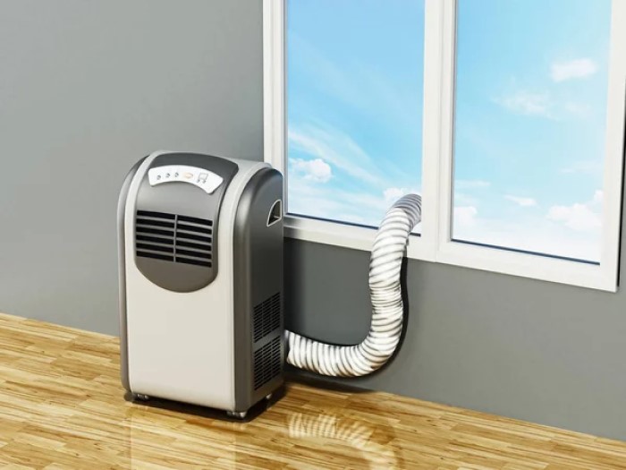 Ilustração realista mostrando um ar condicionado portátil com tubo de ventilação instalado no vidro de janela fechada. O aparelho é branco e prata escuro, lá fora o céu é azul. A parede é cinza e o piso é laminado, cor bege escuro.