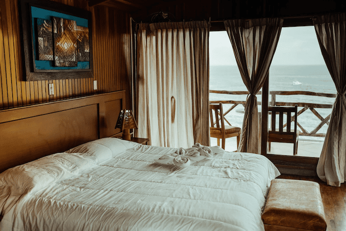 Cama box queen em quarto de madeira com vista para praia. A cama é vista da lateral esquerda e está coberta com um lençol branco, as portas da sacada estão abertas. Há duas cadeiras de madeira e é possível ver o mar. O dia está nublado.