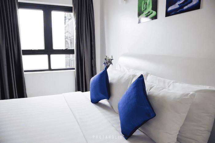 Cama box queen vista de lado um quarto bem iluminado. A roupa de cama é toda branca, exceto por duas almofadas azuis. Do lado esquerdo há uma janela pequena com uma cortina roxa aberta.