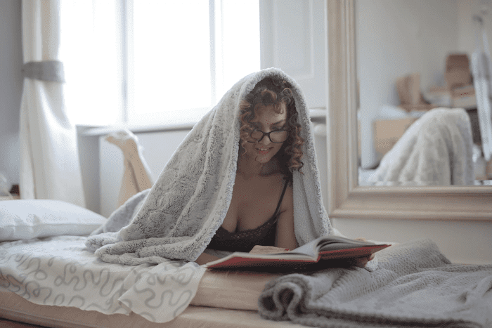Mulher lendo um livro debaixo de uma manta branca sobre uma cama de solteiro. Ela é branca com cabelos loiros cacheados e curtos. Atrás dela há um espelho e ao lado do espelho uma janela com cortina puxada para o lado e que deixa entrar luz.