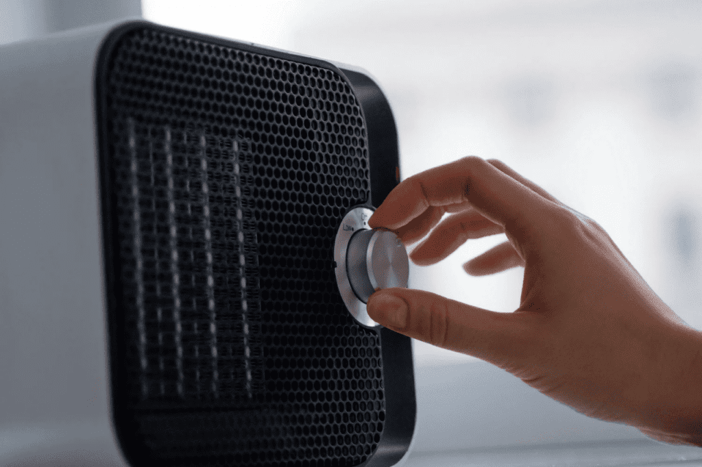 Detalhe mostrando a mão de uma pessoa programando um pequeno climatizador de ar com um botão giratório. O aparelho é preto na parte da frente e branco nas laterais. A mão tem pele clara.