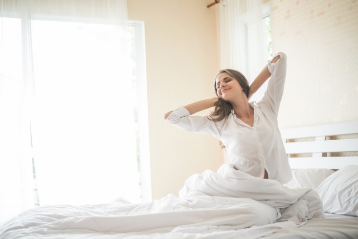 Jovem mulher de cabelos castanhos longos e pele clara, usando camisa branca, se espreguiça com as mãos na nuca em cama de casal e sorri. As roupas de cama são brancas.