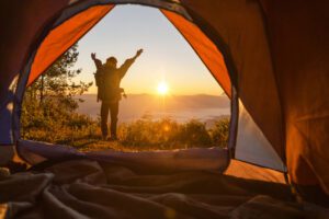 Silhueta de jovem de costas olhando para o nascer do sol com sua barraca de acampamento aberta atrás dele.