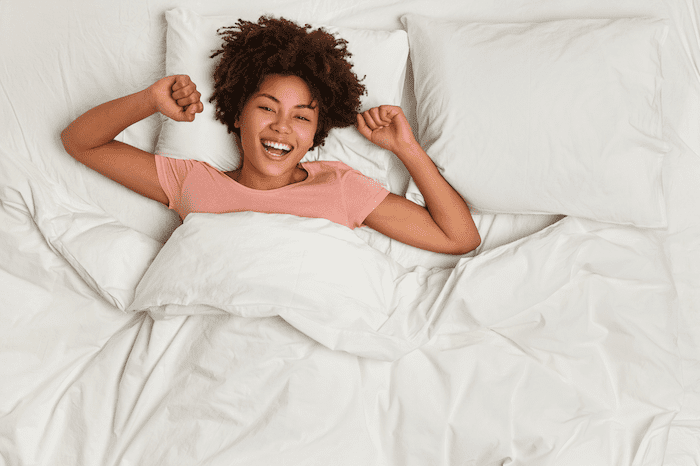 Uma mulher negra jovem, da cintura para cima, vestida com uma blusa rosa, com cabelos cacheados curtos, se espreguiçando e sorrindo, deitada de forma muito confortável sobre uma cama com travesseiros e lençóis brancos.