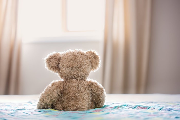 Pequeno urso de pelúcia marrom-claro, meio dourado, colocado de costas, sentado sobre cama com coberta azul. O fundo está desfocado.