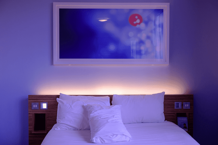 Cama de casal de quarto de hotel fotografada com um ângulo levemente superior e de frente, em quarto iluminado por uma luz azul aconchegante. As roupas de cama são brancas e a parede está toda azulada. A cabeceira da cama é de madeira e tem botões. Há um quadro com pintura abstrata azul e branca na parede atrás da cama.