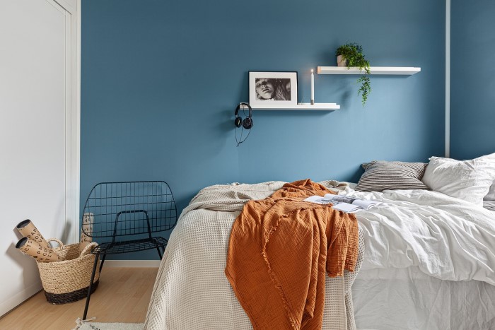 Cama de casal meio desarrumada vista de lado, com lençol e travesseiros brancos, e uma colcha marrom. A parede atrás é azul, e a parede aos pés da cama é branca. O piso é de madeira clara.