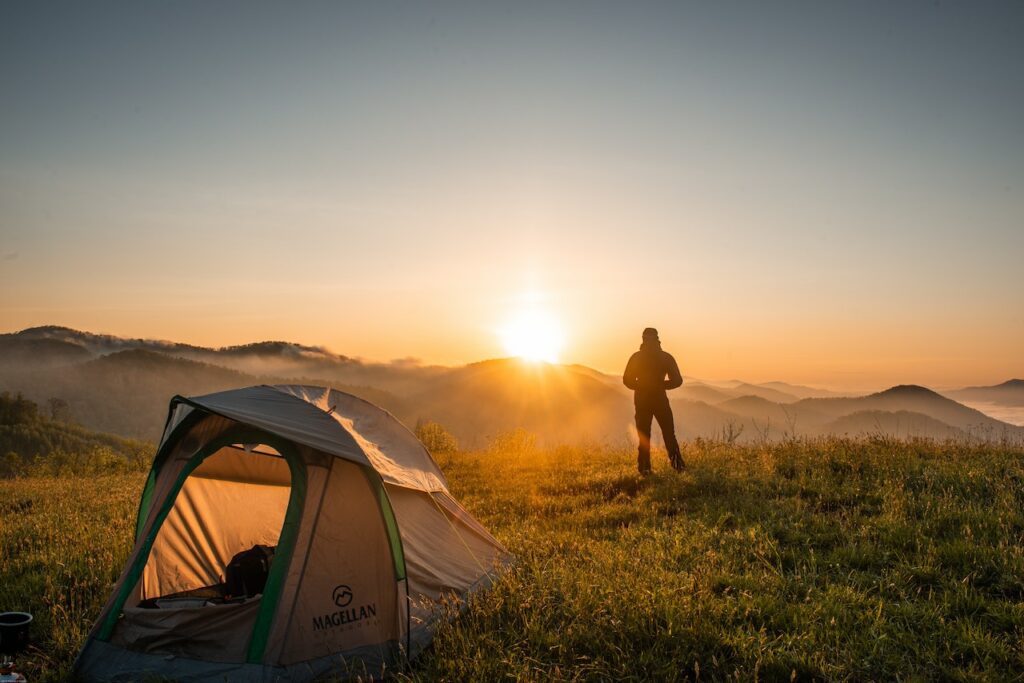 Campista parado do lado de fora da barraca de camping de costas para imagem. É um homem que assiste ao nascer do sol sobre uma colina coberta com grama. A frente dele há uma uma série de montanhas cobertas por neblina. Parece frio.