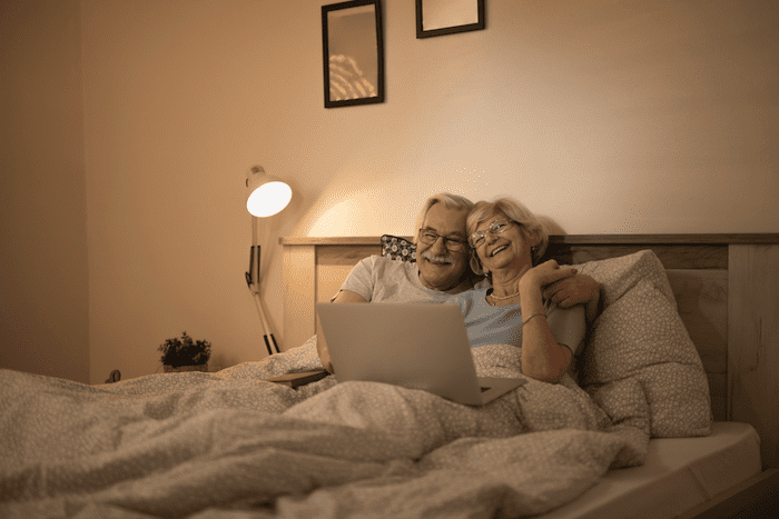 Casal de velhinhos deitados em uma cama, felizes, assistindo em um laptop.