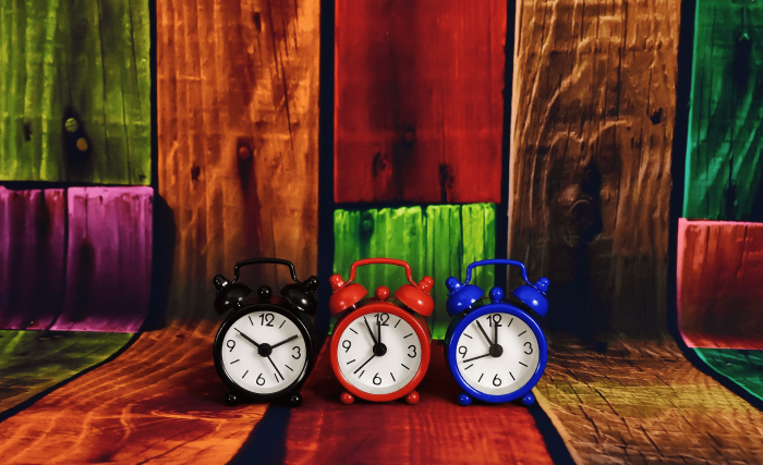 Imagem mostrado 3 despertadores analógicos lado a lado, o primeiro é preto, o segundo é vermelho e o terceiro é azul. Os despertadores estão posicionados sobre uma superfície feita com edição gráfica e que representa várias tábuas de madeiras, rusticamente pregadas, nas cores azul, vermelho, rosa, tons de marrom madeira, e verde. As cores são vibrantes.