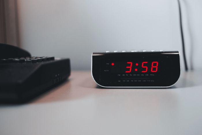 Despertador digital preto, com números vermelhos, marcando 3h e 58 minutos. O despertador está sobre uma mesa branca, ao lado de um aparelho de telefone preto. A parede atrás da mesa é da mesma cor.