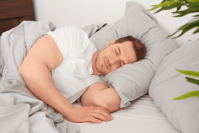 Homem de pele branca e cabelos castanhos deitado e dormindo em uma cama com lençóis cinzas e travesseiros com fronhas cinzas.