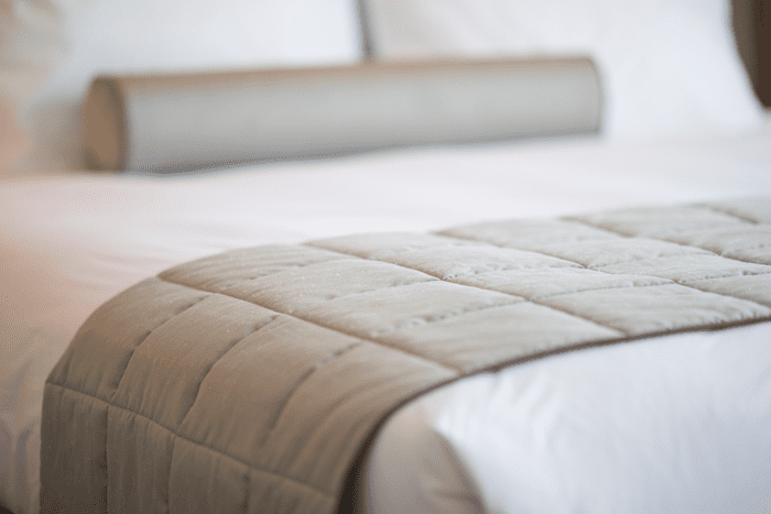 Colchão branco com lençol branco e travesseiros brancos visto de perto. Um adorno esverdeado e um travesseiro de corpo esverdeado são vistos em cima da cama.