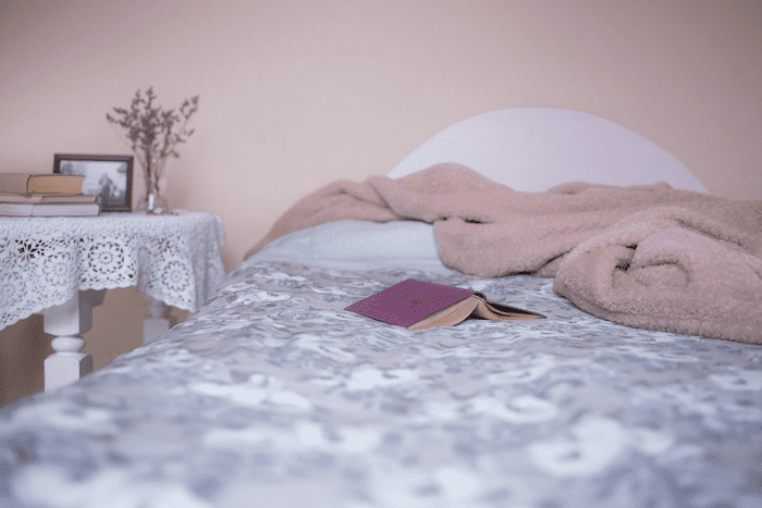 Cama de casal parcialmente vista com lençol de estampas cinzas e coberta rosada. Um livro de capa roxa está aberto em cima da cama.