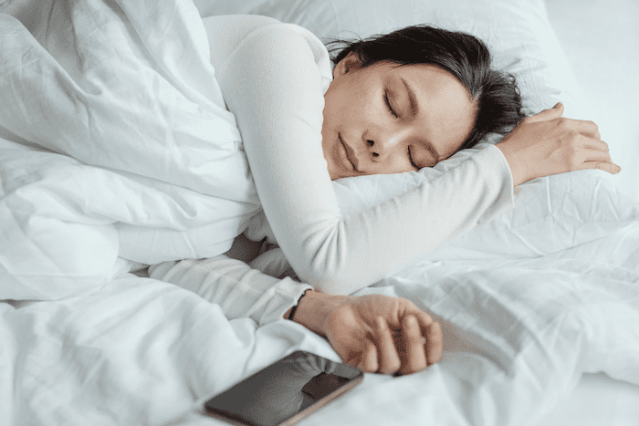 Mulher asiática de cabelos negros, usando blusa de manga comprida branca, dorme tranquilamente enrolada em edredom branco sobre cama de casal. Ela está deitada de lado, com o celular perto do seu corpo.
