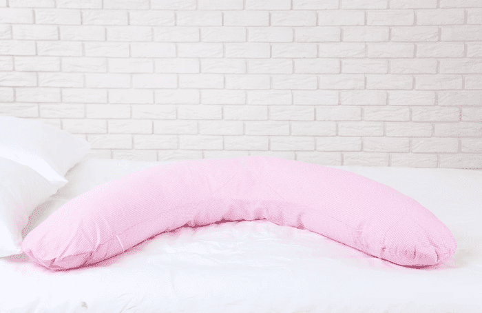Travesseiro de corpo formato retangular levemente curvo em forma de ser um círculo, cor-de-rosa claro, sobre cama de casal com lençol branco. A cama é vista de lado e há dois travesseiros brancos normais na cabeceira. Já a parede é de tijolos aparentes pintados de branco.